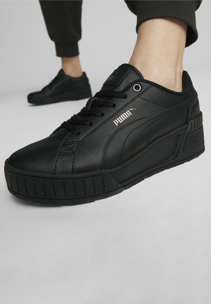 Gros plan d'une personne portant des baskets Puma noires avec des semelles épaisses et des lacets noirs sur un sol gris clair.