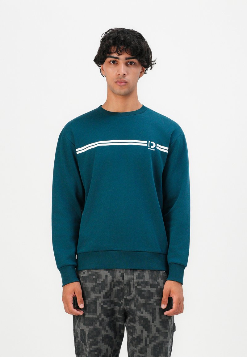 Jeune homme aux cheveux bouclés courts, portant un sweat-shirt teal avec des rayures blanches et un pantalon à motif pixelisé gris, se tenant contre un fond uni.