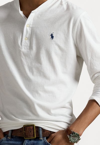 Λευκό μακρυμάνικο πουκάμισο henley με γιακά δύο κουμπιών. Διαθέτει ένα μικρό μπλε λογότυπο στο στήθος. Φοριέται με καφέ ζώνη και πράσινο ρολόι.