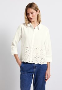 Blusa bianca in cotone con chiusura a bottoni e dettagli ricamati a giorno sull'orlo. Presenta maniche lunghe e una vestibilità morbida.