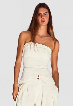 Hosszú barna hajú nő, fehér pánt nélküli felsőt és bő, fehér nadrágot visel, zsebre tett kézzel áll egyszínű háttér előtt.