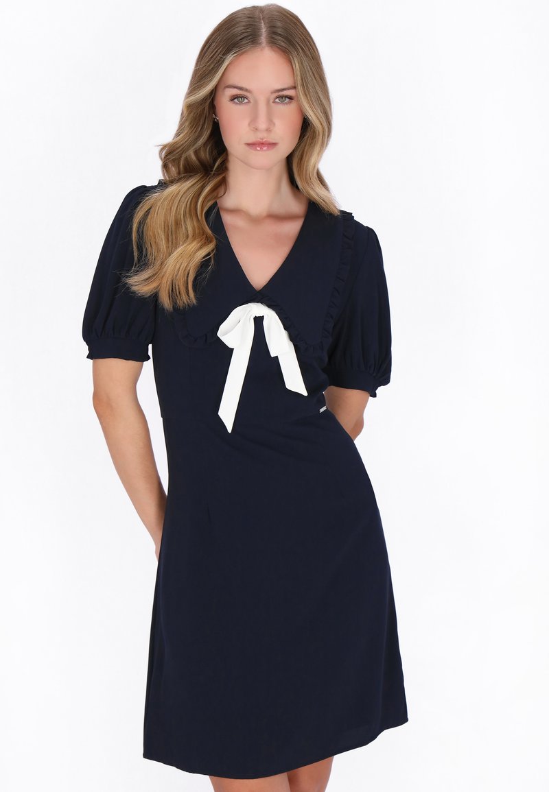 Jonge vrouw met lang blond haar draagt een marineblauwe jurk met pofmouwen en een witte strik bij de grote kraag, staand tegen een witte achtergrond.