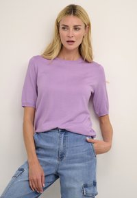 Top corto a maniche corte di colore viola chiaro con scollatura rotonda, realizzato in un tessuto morbido; indossato con jeans in denim blu a vita alta con tasche.