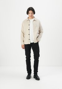 Light beige jacket with buttons, two front pockets, worn over a white shirt. Paired with black jeans and black shoes. Smooth texture.