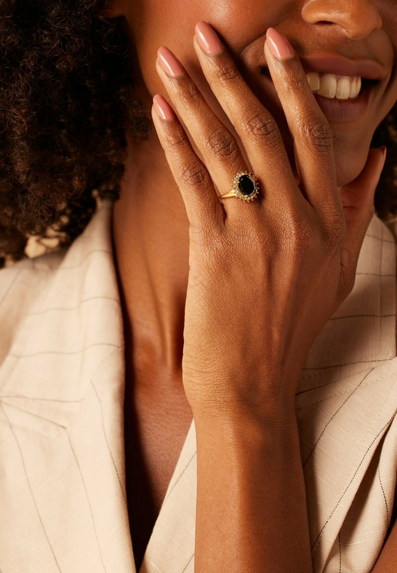 Gouden ring met een ovale zwarte steen omringd door kleine witte accenten, gedragen op een hand die rust tegen een beige stoffen achtergrond.