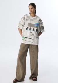 Grafika oversize t-shirt w beżowym kolorze z różnymi monochromatycznymi wzorami, niebieskimi i czerwonymi akcentami, w połączeniu z khaki, teksturowanymi spodniami typu wide-leg.
