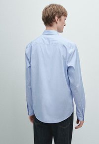 Camicia a righe azzurro chiaro con colletto button-down, maniche lunghe e hem curvo. Il tessuto è liscio con una leggera lucentezza.