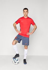 T-shirt Nike rouge sportif avec des accents noirs, associé à un short bleu arborant un logo orange. Le modèle porte des chaussettes blanches et des crampons noirs, se tenant debout sur un ballon de football.