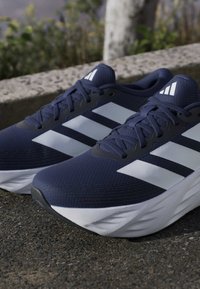 Pair of navy blue Adidas running shoes with white stripes and white midsoles placed on asphalt near a stone curb.