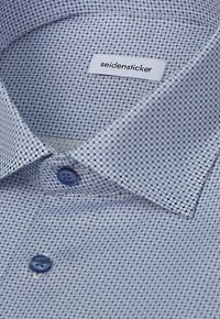 Gros plan d’une chemise habillée pliée à motifs avec des boutons bleus et une étiquette blanche indiquant le nom de la marque Seidensticker à l’intérieur du col.