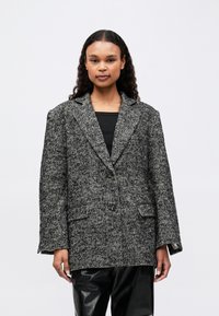 Mladá žena s dlhými kučeravými vlasmi, oblečená v šedom oversize blejzri s herringbone vzorom a čiernych kožených nohaviciach, stojí pred jednoduchým pozadím.