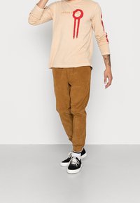Beige pitkähihaisessa paidassa on punainen graafinen kuvio ja teksti, ja se on yhdistetty vaaleanruskeisiin corduroypantoihin sekä mustiin lenkkkenkiin, joissa on valkoisia yksityiskohtia.