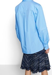 Light blue long-sleeve shirt with a button-down collar and cuffs, paired with a textured navy skirt featuring gold accents and fringed hem.