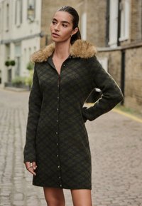 Cappotto a quadri verde e blu navy fino al ginocchio, con colletto in pelliccia sintetica, chiusura con bottoni e maniche lunghe. Tessuto morbido e lavorato a maglia dalla texture soffice.
