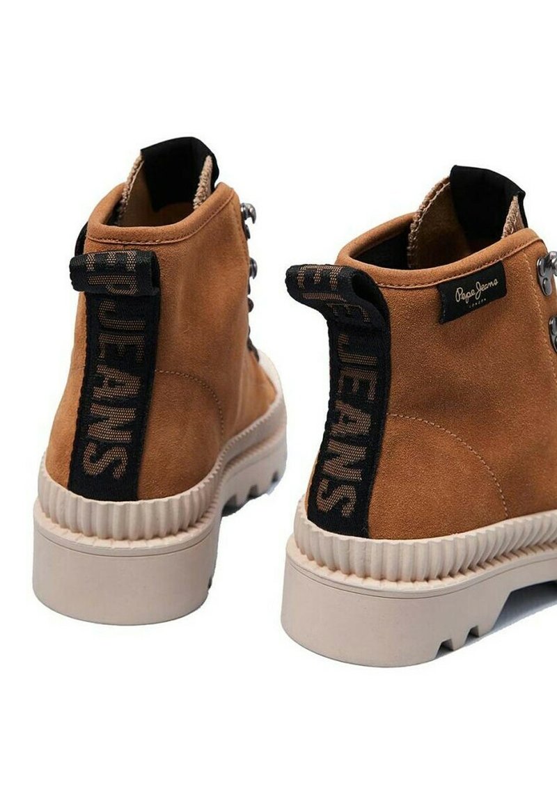 Botines de ante marrón con una superficie texturizada, que presentan una lengüeta de tela negra en el talón etiquetada como "JEANS" y una suela gruesa de goma blanca.