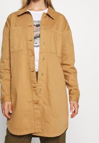 Beige bomulls skjortjacka med krage, två bröstfickor och rak skärning. Har stora runda knappar och en slät textur.