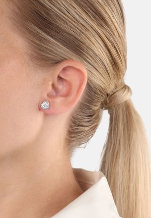 Primer plano de la oreja de una mujer con un pendiente de plata en forma de corazón y el cabello rubio recogido en una coleta.