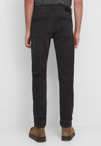 Hombre con jeans slim-fit negros y botas de ante marrones, de pie contra un fondo gris claro liso, visto desde la parte de atrás.
