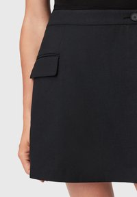 Zwarte rok van gladde stof. Bevat een zijzak met een klep. Strakke lijnen en een ontworpen stijl, de zoom eindigt net boven de knie.