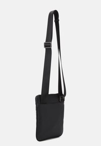 Černá kabelka přes rameno s texturovaným povrchem, nastavitelným popruhem, zipovým uzávěrem a minimalistickým designem. Má plochý, obdélníkový tvar.