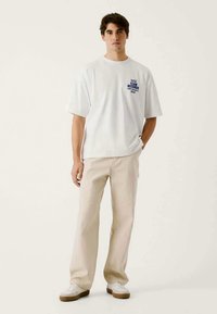 Balta kokvilnas t-kreklā ar zilu grafisko tekstu uz priekšpuses, apvienota ar gaiši bēšām biksēm un baltām čībām, stāvot uz neitrāla fona.