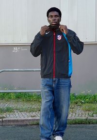 Sort vindjakke med en rød lynlås og blå detaljer, med et logo på venstre bryst. Bæres med blå jeans og hvide sneakers.