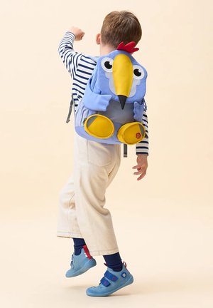 Child wearing striped shirt, light pants, and blue shoes jumps, carrying a blue and yellow bird-shaped backpack with red crest.