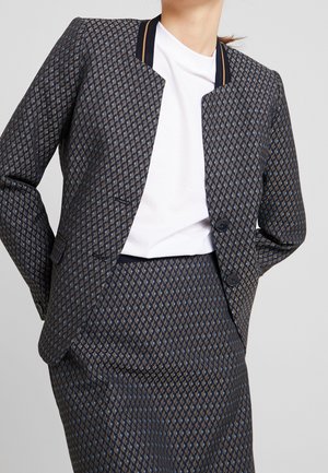 Femme portant un blazer à motif à deux boutons et une jupe assortie par-dessus un t-shirt blanc à col rond, se tenant avec les mains derrière le dos.