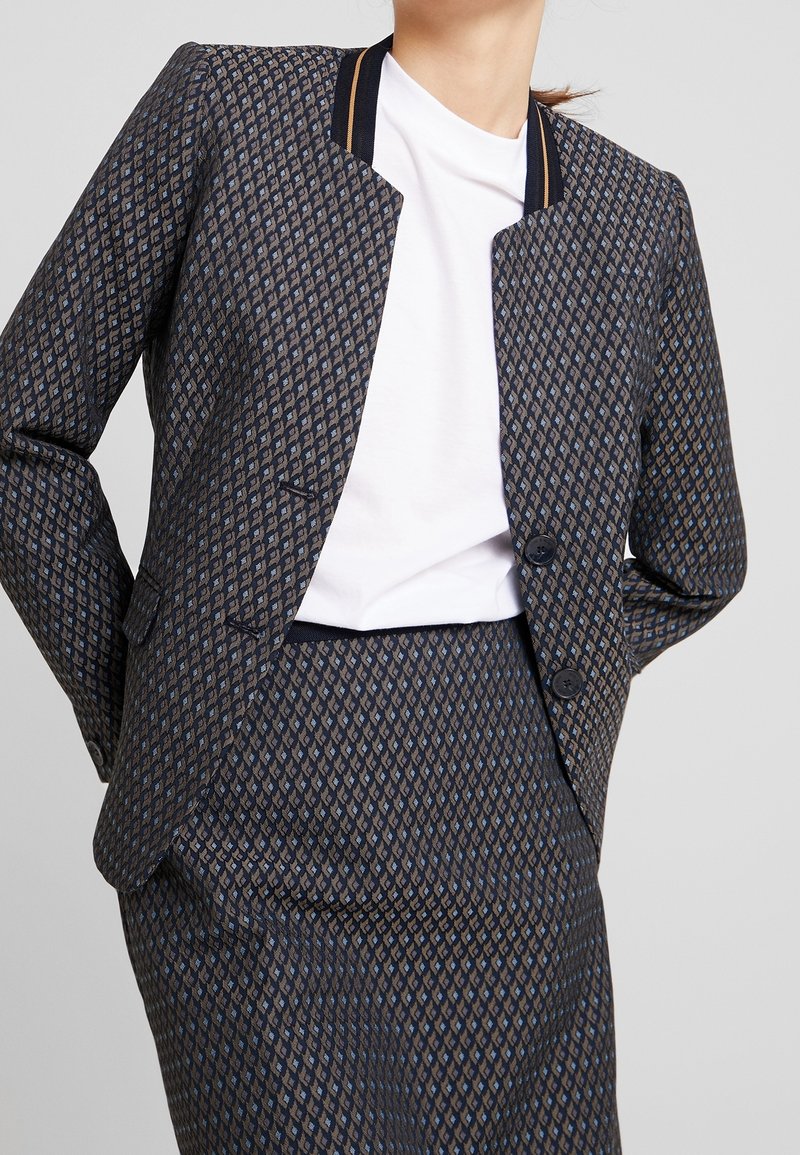 Femme portant un blazer à motif à deux boutons et une jupe assortie par-dessus un t-shirt blanc à col rond, se tenant avec les mains derrière le dos.