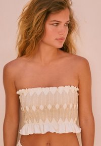 Jauna sieviete ar viļņainiem blondiem matiem, tērpusies baltā strapless augšdaļā ar bēšām zigzaga rakstiem un volānu malām, skatās sāniski.