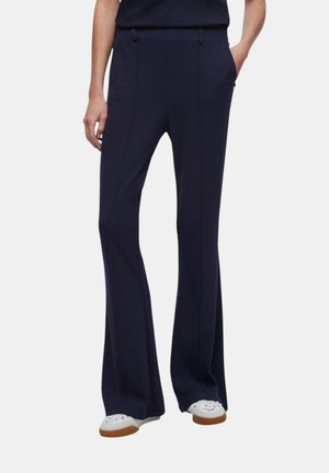 Pantaloni blu navy su misura a gamba larga indossati con scarpe da ginnastica bianche, modello in piedi con una mano in tasca su uno sfondo semplice.
