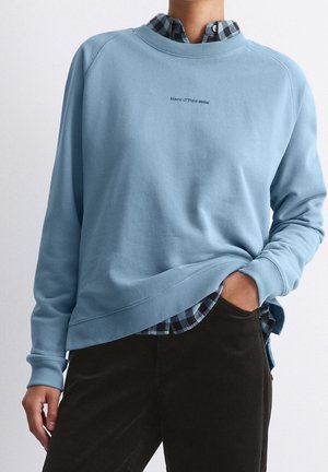 Personne portant un sweat-shirt bleu clair sur une chemise à carreaux noire et bleue, la main dans la poche d'un pantalon foncé.