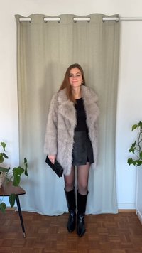 Jasnoszary płaszcz z futra ekologicznego z szerokim kołnierzem, czarna bluzka, skórzana mini spódnica, czarne rajstopy i czarne, sięgające kolan, szpiczaste botki, trzymająca czarną kopertówkę.