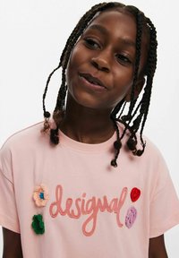 Ljusrosa bomulls-t-shirt med broderad "desigual" text i kursiv, accentuerad med färgglada blommor och prickar. Korta ärmar.