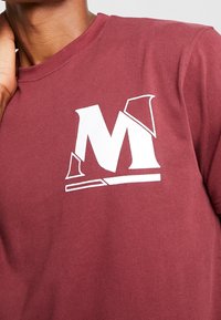 Bordeaux farvet bomulds T-shirt med et stort hvidt grafisk bogstav "M" med vinklede udsparinger, korte ærmer og rund halsudskæring.