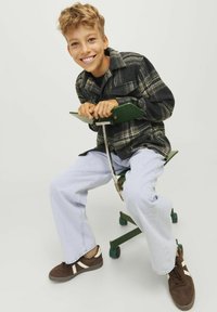 Camisa de franela a cuadros en negro y verde, pantalones vaqueros holgados en azul claro y zapatillas marrones con detalles en blanco; sentado en un taburete rodante verde.