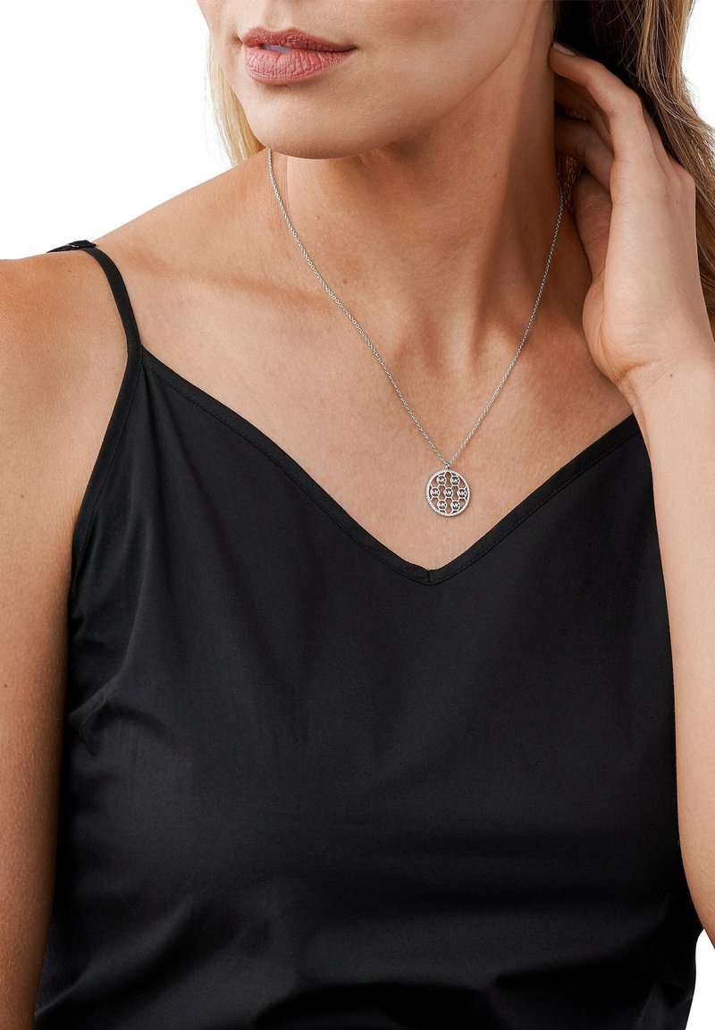 Femme portant un haut noir à fines bretelles et un collier argenté avec un pendentif circulaire, touchant ses cheveux près du cou.