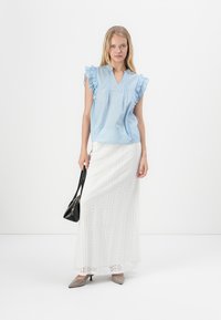 Blusa de manga corta azul claro con bordes rizados, combinada con una falda maxi blanca de encaje con un patrón floral. Accesorios incluyen un bolso negro.