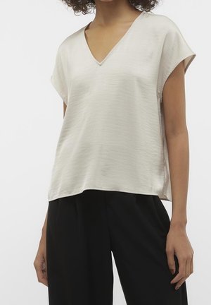 Woman wearing a short-sleeve, light beige V-neck blouse with loose black pants against a plain background.