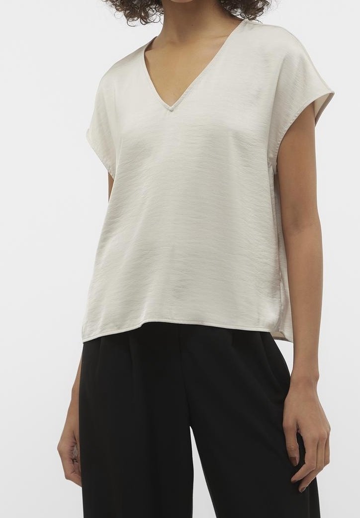 Femme portant un chemisier beige clair à manches courtes avec un col en V et un pantalon noir ample sur un fond uni.