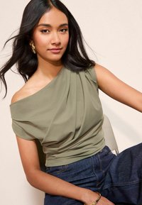 Olive rohelise tooniga õlgadelt avatud top, pehme kangas ja tekstuurilised voldid, millele on lisatud tume denimiseer. 