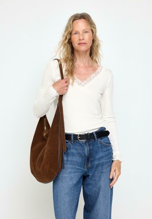 Kvinde med krøllet blondt hår iført en hvid blondebeklædt top og blå jeans, der holder en stor brun ruskind skuldertaske.