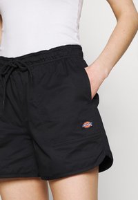 Černé šortky vyrobené z tkané látky, s elastickým pasem, šňůrkou, bočními kapsami a nášivkou s logem na levé straně.