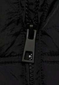 Sectiune a unei jachete negre cu fermoar, prezentând un material texturat, un fermoar negru proeminent și o mânere rectangulară pentru o prindere ușoară.