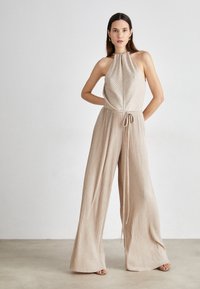 Beige pleated jumpsuit with a halter neck, tie waist, and wide-leg design. Smooth, textured fabric with vertical stripes.
