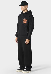 Černá mikina s kapucí s oranžovým designem listu na kapse na prsou, spárovaná s černými tepláky a černými teniskami. Uvolněný střih a materiál z bavlny.