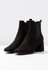 Botas de ante negras hasta el tobillo con punta afilada y tacón de bloque. Presentan paneles laterales elásticos para facilitar el uso. Textura suave, diseño minimalista.