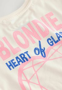 Kermanvärinen puuvillapaita, jossa on suuri vaaleanpunainen ja sininen teksti, joka lukee "BLONDIE HEART of GLASS", ja taustalla on geometrinen kuvio.