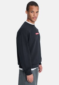 Zwarte sweatshirt met een ronde hals en lange mouwen. Beschikt over een rood grafisch logo aan de voorkant. Zacht materiaal met een relaxte pasvorm.