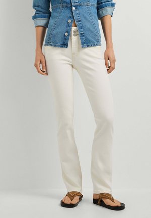 Kvinde iført hvide, højtaljede jeans, en opkrøllet denimjakke og brune sandaler med åbne tæer står mod en ensfarvet baggrund.